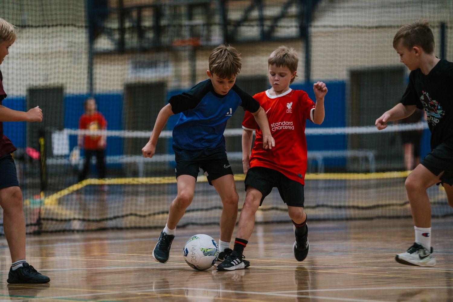 Two young boys closely jostle for a soccer ball on an indoor court, while other children watch from the sides during a casual match.